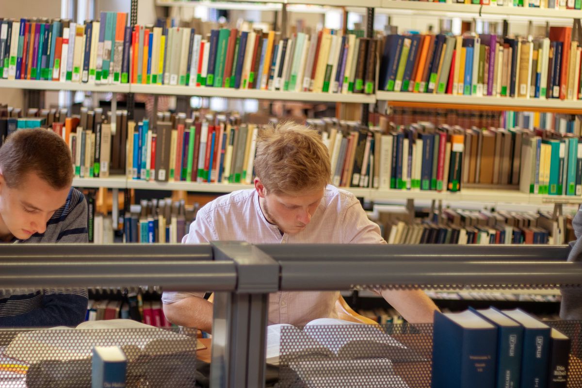 Zwei Studenten sitzen an einem Tisch in der Bibliothek Albertina. Hinter ihnen sind Bücherregale. Auf ihren Tischen sind ebenfalls viele Bücher, in jeweils einem davon lesen die beiden Studierenden konzentriert. Durch die vielen bunten Buchrücken ist das Foto sehr farbenfroh.