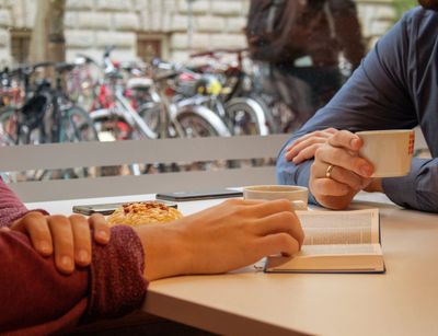 In der Cafeteria des Studentenwerks im Geisteswissenschaftlichen Zentrum der Uni Leipzig sitzen zwei Stunden an einem Tisch und trinken Kaffee. Man sieht keine Gesichter. Der linke von beiden hat seine rechte Hand in einer geöffneten Bibel liegen. Durch das Fenster hinter ihnen sieht man einen Teil der Mauer der gegenüberliegenden Bibliothek Albertina. Außerdem stehen draußen einige Fahrräder an Fahrradbügeln.