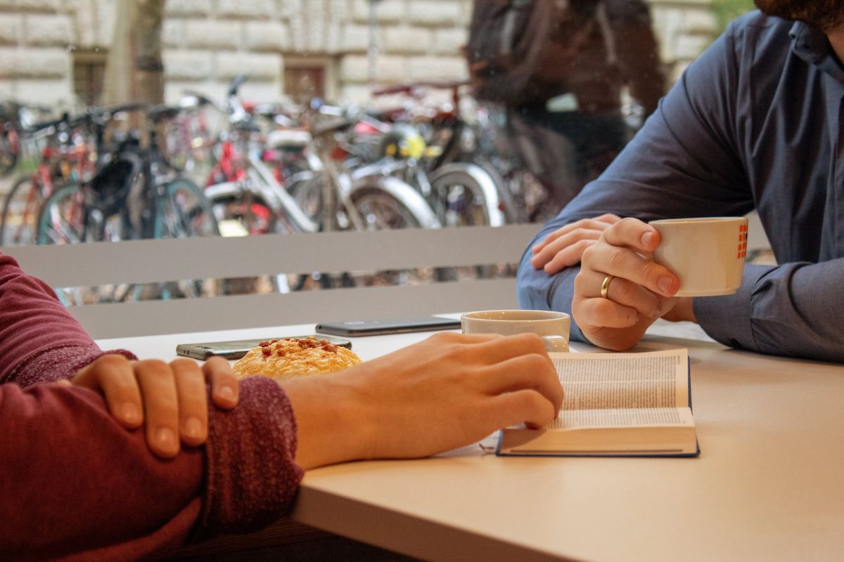 zur Vergrößerungsansicht des Bildes: In der Cafeteria des Studentenwerks im Geisteswissenschaftlichen Zentrum der Uni Leipzig sitzen zwei Stunden an einem Tisch und trinken Kaffee. Man sieht keine Gesichter. Der linke von beiden hat seine rechte Hand in einer geöffneten Bibel liegen. Durch das Fenster hinter ihnen sieht man einen Teil der Mauer der gegenüberliegenden Bibliothek Albertina. Außerdem stehen draußen einige Fahrräder an Fahrradbügeln.