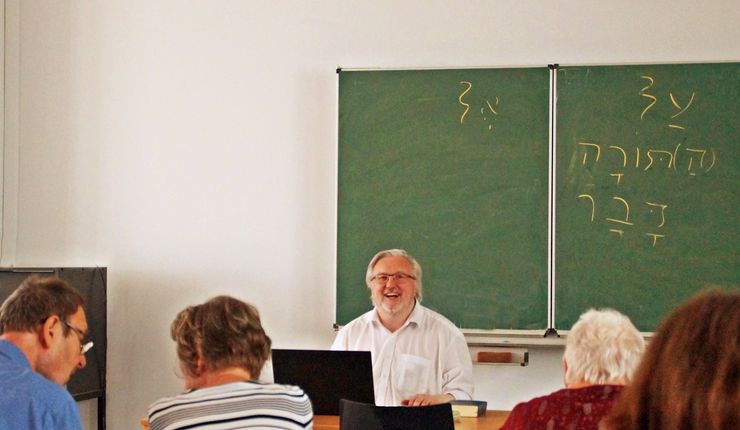 Das Foto ist im Hörsaal der Theologischen Fakultät entstanden. In der Mitte ist der Dozent zu sehen. Er sitzt an einem Tisch, hat ein Notebook vor sich und guckt freudig und erwartungsvoll die Zuhörenden an. Hinter ihm ist eine Kreidetafel. An dieser stehen hebräische Schriftzeichen. Von hinten sieht man einige der Zuhörenden an Tischen sitzen.