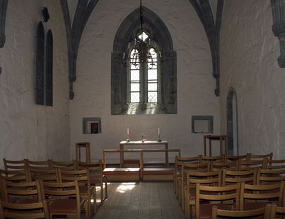 Zu sehen ist eine leere Kirche. Die Kirche ist schlicht, es gibt Holzstühle und einen schlichten Holzaltar. Durch das Fenster hinter dem Altar scheint Sonne herein.