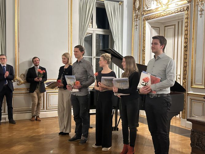 Foto: Junge Absolventinnen und Absolventen stehen mit Mappe und Rose im Alten Senatssaal und genießen den Moment.