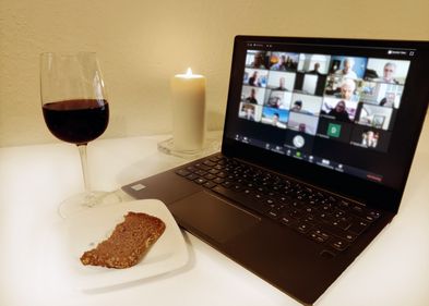 Laptop mit Videokonferenz-Kacheln, angezündete Kerze, Brot und Weinglas