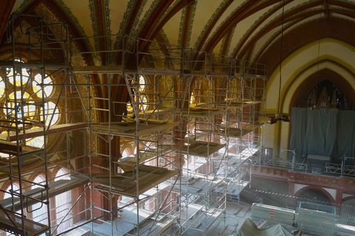 Kircheninnenraum mit leuchtenden Glasfenstern, davor Baugerüste