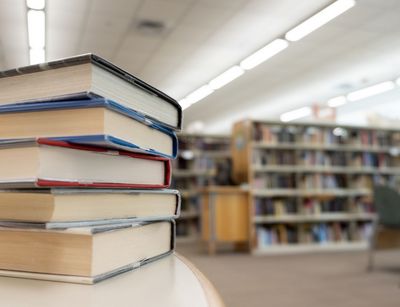 Im Vordergrund: Bücherstapel liegend, im Hintergrund: Bibliotheksregale