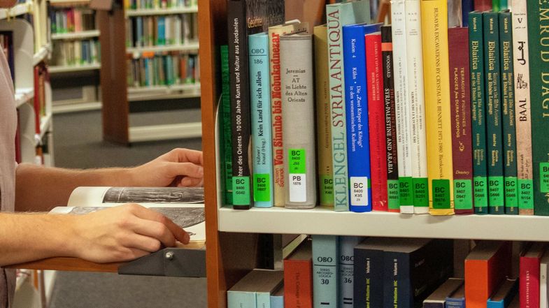 Das Foto ist in der Bibliothek Albertina aufgenommen. Man sieht einige Regale mit vielen Büchern darin. Seitlich an einem Regal kann man eine kleine Holzplatte ausklappen. Auf dieser ausgeklappten Platte liest ein Student stehend in einem Buch.