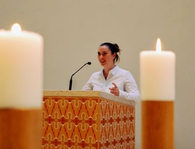 Das Foto wurde in der katholischen Kirche St. Trinitatis in Leipzig aufgenommen. Eine Studentin steht am Pult der Kirche und liest. Links und rechts von ihr sind Kerzen zu sehen, welche sich aber nahe der Kamera befinden und nicht direkt neben ihr stehen.