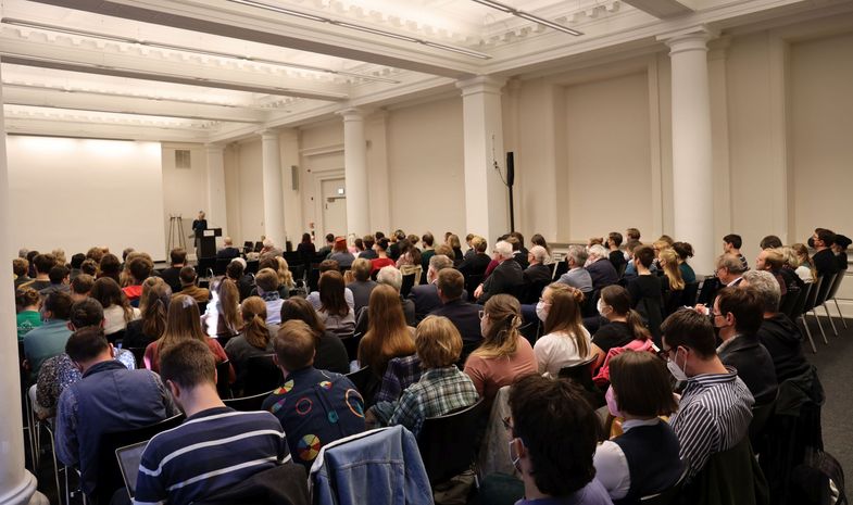 Viele Menschen sitzen in einem großen Raum. Im Hintergrund ist eine Bühne und ein Podium zu sehen. Eine Frau spricht. Foto: Maria Zink.