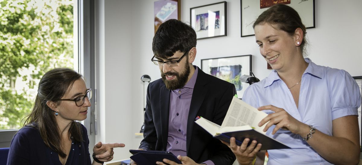 Mitarbeiter:innen des Instituts für Neutestamentliche Wissenschaft, Foto: Andreas Schüle. Zwei Frauen und ein Mann sitzen am und auf einem Schreibtisch. Eine Frau hält ein Buch in der Hand. Die andere zeigt auf das Tablet in den Händen des Mannes.