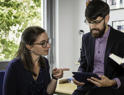 Mitarbeiter:innen des Instituts für Neutestamentliche Wissenschaft, Foto: Andreas Schüle. Zwei Frauen und ein Mann sitzen am und auf einem Schreibtisch. Eine Frau hält ein Buch in der Hand. Die andere zeigt auf das Tablet in den Händen des Mannes.