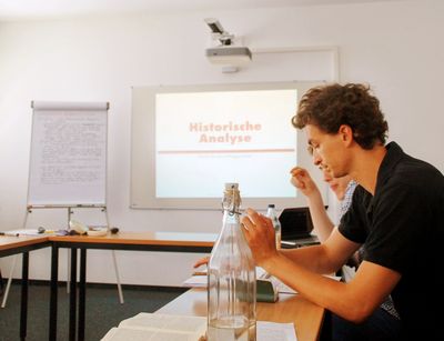 Das Foto ist in einem der Seminarräume in der Theologischen Fakultät entstanden. In der Mitte sieht man die Worte "Historische Analyse" über den Beamer an eine weiße Tafel projiziert. Rechts sitzen Studierenden seitlich zur Tafel. Diese schauen nicht nach vorne, sondern vor sich in ein Buch oder auf ein Blatt Papier.