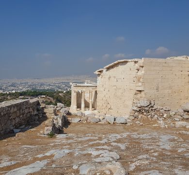 Blick von der Akropolis auf die Stadt Athen, Foto: Nicole Oesterreich. Auf der rechten Seite befinden sich griechische Tempelruinen, links eine Mauer. Im Hintergrund eine Stadt und blauer Himmel mit wenigen Wölkchen.
