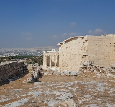 Athen_Akropolis Blick von der Akropolis auf die Stadt Athen, Foto: Nicole Oesterreich. Auf der rechten Seite befinden sich griechische Tempelruinen, links eine Mauer. Im Hintergrund eine Stadt und blauer Himmel mit wenigen Wölkchen.
