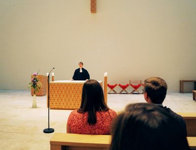 Das Foto wurde in der katholischen Kirche St. Trinitatis in Leipzig aufgenommen. In der Mitte einige Meter entfernt sieht man einen Studierenden im Talar, der hinter dem Pult steht. Er steht mit dem Gesicht zu Gemeinde und Kamera. Rechts im Bild sitzen drei Studierenden in den Bänken, diese sieht man nur von hinten.