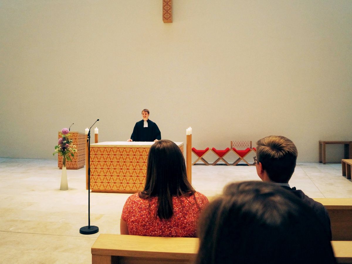 zur Vergrößerungsansicht des Bildes: Das Foto wurde in der katholischen Kirche St. Trinitatis in Leipzig aufgenommen. In der Mitte einige Meter entfernt sieht man einen Studierenden im Talar, der hinter dem Pult steht. Er steht mit dem Gesicht zu Gemeinde und Kamera. Rechts im Bild sitzen drei Studierenden in den Bänken, diese sieht man nur von hinten.