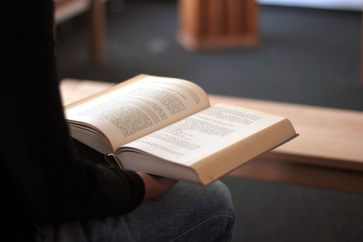 zur Vergrößerungsansicht des Bildes: Eine Studentin sitzt im Andachtsraum der Theologischen Fakultät auf einer Holzbank. Man sieht sie nur verschwommen von der Seite, vor allem den rechten Arm und das rechte Bein. In der Hand hält sie ein dickes theologisches Buch, in welchem sie list. 