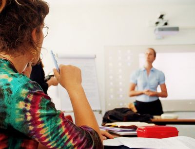 Das Foto ist in einem Seminarraum an der Theologischen Fakultät entstanden. Links im Foto sieht man scharfgestellt eine Studentin mit buntem Oberteil von der Seite. Auf dem Tisch vor ihr liegt Papier, außerdem eine rote Brotdose. Rechts im Bild sieht man unscharf die Dozentin. Sie hört gerade einem der Studierenden zu. Hinter ihr hängt eine weiße Tafel, auf welche etwas mit dem Beamer projiziert wird.