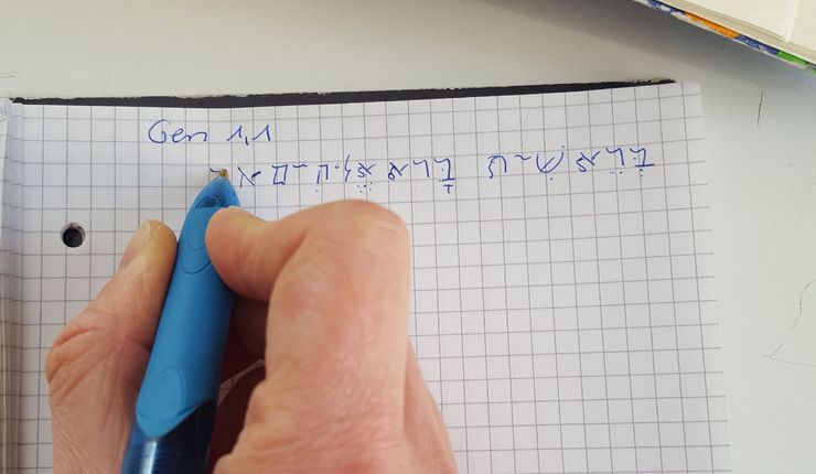 Eine Hand schreibt den ersten Vers der Bibel auf Hebräisch.