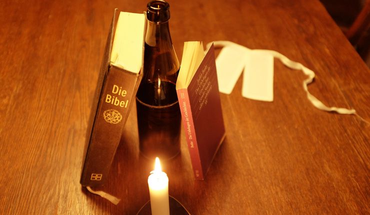 Auf dem Foto zu sehen sind Gegenstände auf einem Holztisch, welche nur schwach von einer Kerze beleuchtet werden. In der Mitte steht eine Bierflasche. Daran angelehnt sind eine Lutherbibel und ein Buch von Judith Butler. Im Hintergrund liegt ein Beffchen.