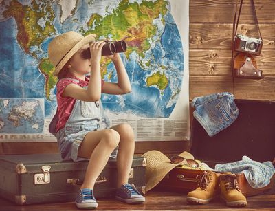 Ein Mädchen sitzt vor einer großer Weltkarte. Sie guckt durch ein Fernglas, neben ihr liegen verschiedene und sitzt auf einem Koffer. Neben ihr liegt ein weiterer Koffer mit Dingen, die man für eine Forschungsreise gebrauchen kann.