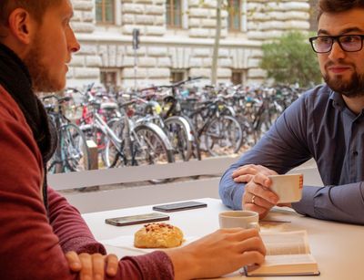 In der Cafeteria des Studentenwerks im Geisteswissenschaftlichen Zentrum der Uni Leipzig sitzen zwei Stunden an einem Tisch und trinken Kaffee. Auf dem Tisch liegt eine Bibel. Durch das Fenster hinter ihnen sieht man einen Teil der Mauer der gegenüberliegenden Bibliothek Albertina.