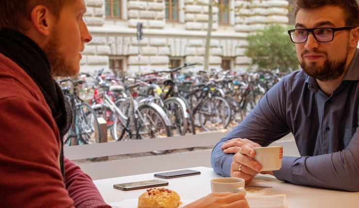 In der Cafeteria des Studentenwerks im Geisteswissenschaftlichen Zentrum der Uni Leipzig sitzen zwei Stunden an einem Tisch und trinken Kaffee. Auf dem Tisch liegt eine Bibel. Durch das Fenster hinter ihnen sieht man einen Teil der Mauer der gegenüberliegenden Bibliothek Albertina.