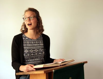 Eine Studentin steht an einem Pult aus Holz im Andachtsraum der Fakultät. Sie spricht gerade, weshalb ihr Mund geöffnet ist. Ihr Hände halten die auf dem Pult aufliegende aufgeklappte rote Bibel.