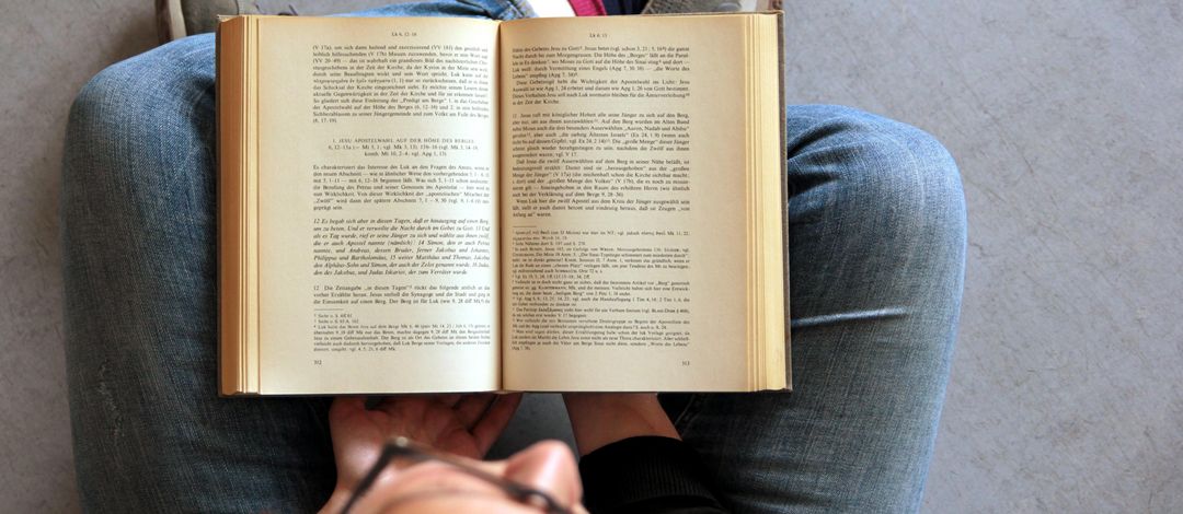 Eine Studentin sitzt auf einem grauen Boden im Schneidersitz. Das Foto ist von oben aufgenommen. In ihrem Schoß liegt ein aufgeschlagenes dickes theologisches Buch. Es handelt sich dabei um einen Kommentar zum Lukasevangelium. 