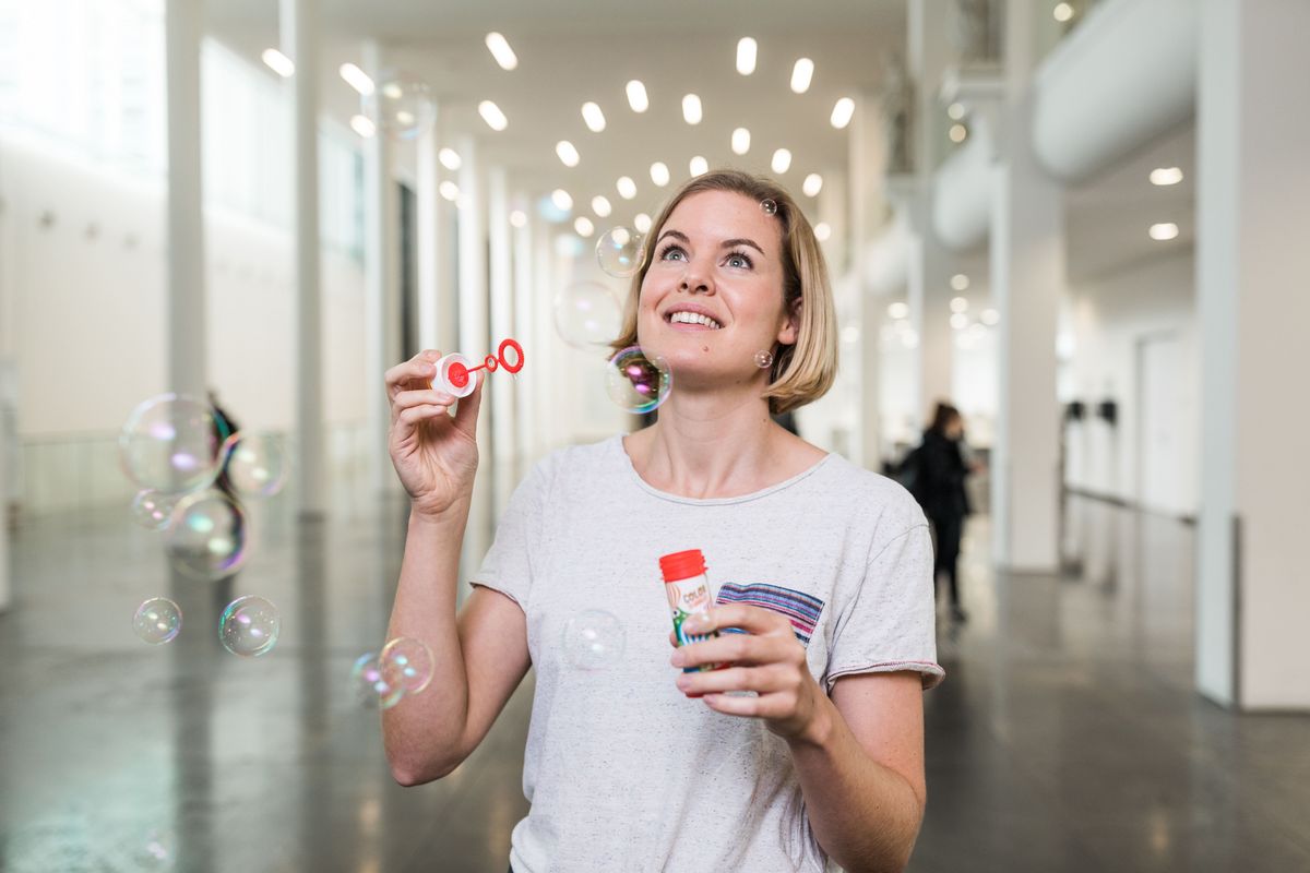 zur Vergrößerungsansicht des Bildes: Eine Frau in einem großen Raum, die Seifenblasen pustet. Bildunterschrift: Studienstart, Foto: Christian Hüller.