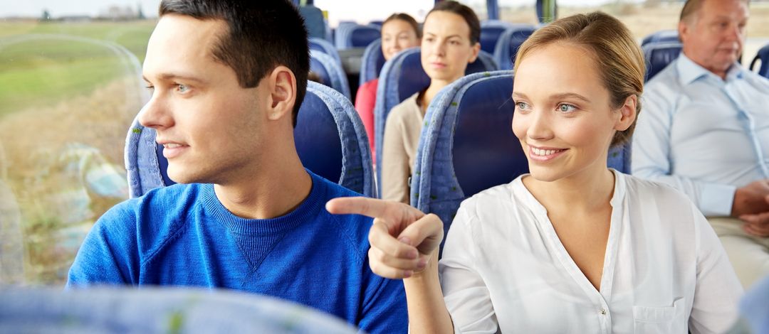 Personen sitzen in einem Bus. Eine Frau zeigt ihrem Sitznachbarn etwas draußen.