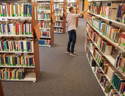 Zu sehen ist ein Gang in der Bibliothek Albertina mit vielen hohen und vollen Bücherregalen. Viele farbige Buchrücken machen das Foto sehr bunt. In der Mitte nimmt sich ein Student ein Buch aus einem der Regale.