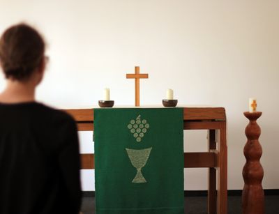 In der Mitte steht der hölzerne Altar im Andachtsraum der Fakultät. Auf ihm steht ein goldenes Kreuz. Eine grüne Altardecke ist mit einem silbernem Kelch und Trauben bestickt. Links im Bild sitzt eine Studentin, der Kamera mit dem Rücken zugewandt. Sie ist nur unscharf zu sehen.