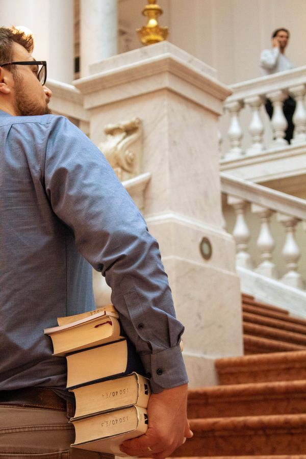 Ein Student geht auf der große Haupttreppe in der Bibliothek Albertina nach oben. Man sieht ihn schräg rechts von hinten. Er trägt eine Brille und guckt nach oben. Unter seinem rechten Arm sind vier dicke Bücher.