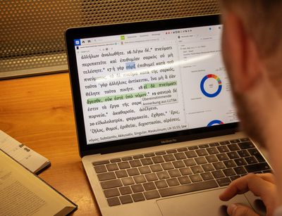 Das Foto guckt einem Studierenden über die Schulter. Dieser sitzt in der Bibliothek Albertina an einem Tisch und hat sein MacBook Pro geöffnet. Das Programm logos ist geöffnet, mit welchem sich der Studierende den griechischen Text des Neuen Testaments anguckt.