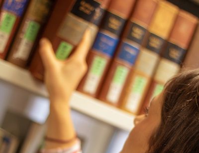 Eine Studentin nimmt sich ein großes und dickes Buch aus einem Regal in der Bibliothek Albertina. Man sieht sie dabei von der Schulter an und schräg von hinten Links.