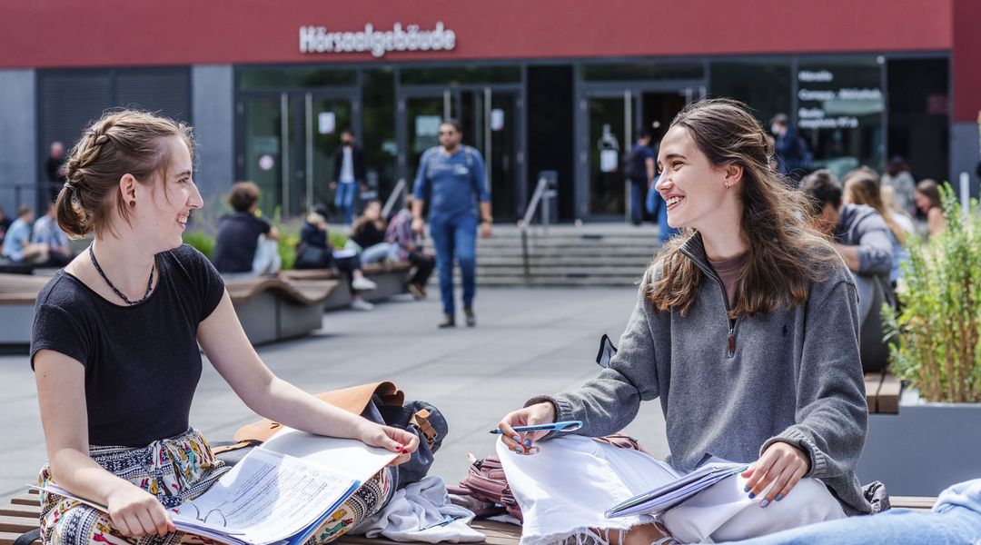 Zwei Studentinnen sitzen auf dem Uni-Innenhof im Schneidersitz auf einer Bank und unterhalten sich. Im Hintergrund sind unscharf Menschen zu sehen, die ins Hörsaalgebäude gehen.